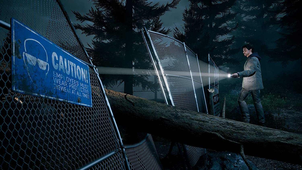 Alan Wake Remastered game scene showing a male character with a flashlight near a fallen tree and chain-link fence at night.